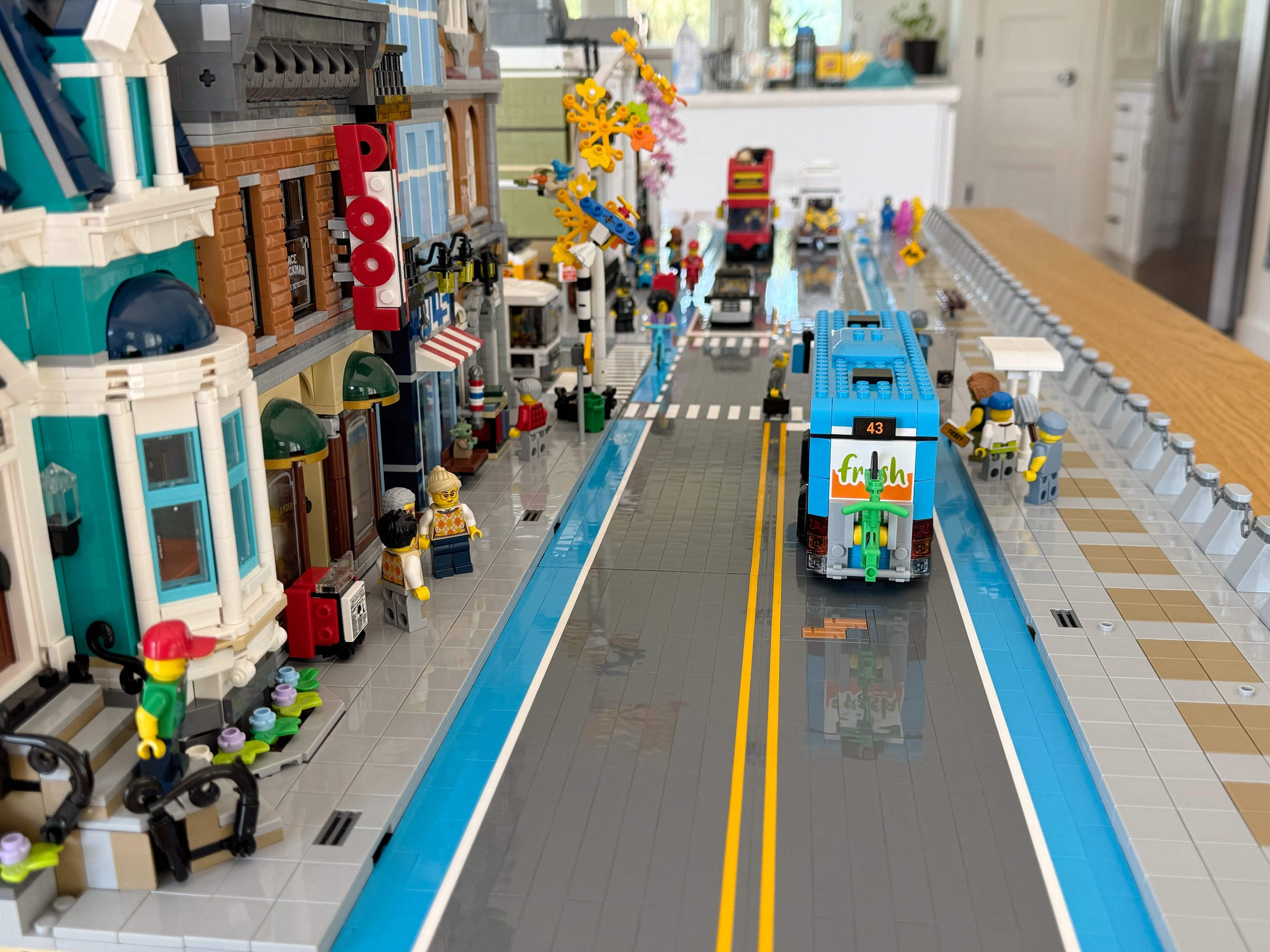 A look down the Main Street of a LEGO city with buildings on the left. An azure bus sits at a bus stop in the right lane of the street